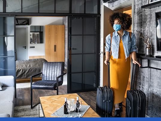 Woman wearing face mask entering vacation rental with luggage