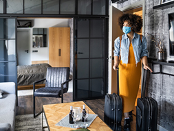 Woman wearing face mask entering holiday rental with luggage